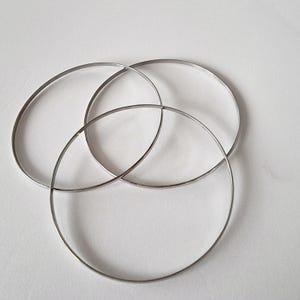 May include: Three silver-coloured, thin metal bangles are interlocked, creating a circular design. The bangles are smooth and reflective, with a simple, minimalist aesthetic. The jewellery is set against a plain white background.
