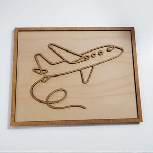 May include: Wooden wall art featuring a detailed outline of an airplane. The airplane is depicted in a side view with its wings, tail, and a looping trail. The artwork is set within a rectangular wooden frame.