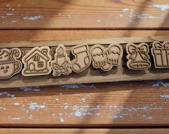 May include: A wooden tray displays seven laser-cut Christmas shapes. These include a mug, house, candle, stocking, mittens, bell, and gift. The shapes are light brown against a wooden background.