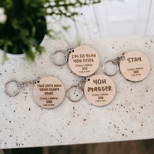 May include: A collection of wooden keychains with engraved messages and silver keyrings. The keychains feature phrases like "This isn't how your story ends," "I'm so glad you exist," "You matter," and "Stay." Each keychain includes the Crisis Lifeline number 988.
