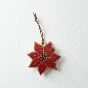 Könnte beinhalten: Weihnachtsbaumschmuck aus Holz in Form eines Weihnachtssterns mit rotem Blumendesign und brauner Lederschnur zum Aufhängen. Die Blume hat einen natürlichen Holzrand und eine schwarze Mitte. Eine festliche Weihnachtsdekoration.