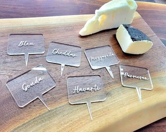 May include: Six clear acrylic cheese markers with black lettering on a wooden cutting board. The markers are labeled: Bleu, Cheddar, Mozzarella, Gouda, Havarti, and Parmesan.