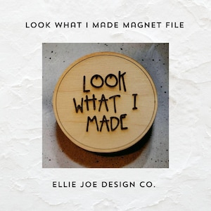 May include: A round wooden magnet with the text "LOOK WHAT I MADE" in black lettering. The magnet is on a light brown surface.