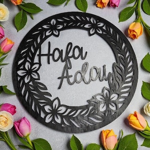 May include: A black wooden circular wall decor with the names "Hafa Adai" cut out in a cursive font. The circular frame is decorated with floral and leaf cutouts. The background is light gray, with colorful flowers and green leaves.