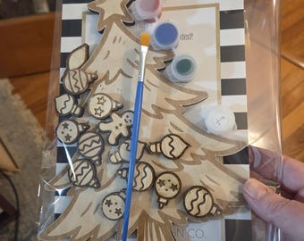 Puede incluir: Un kit de bricolaje de árbol de Navidad en un paquete de plástico transparente. El kit incluye un árbol de madera, adornos, botes de pintura en rojo, azul y verde, un pincel y un pequeño recipiente de pegamento. El fondo presenta un diseño a rayas blancas y negras.