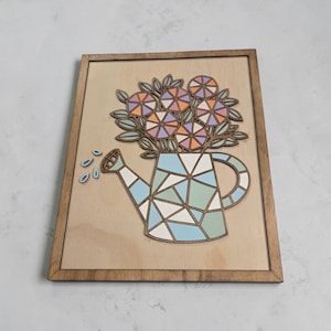 May include: Wooden framed wall art depicting a watering can design with a bouquet of flowers. The watering can has geometric shapes in blue, green, and white. The flowers are in shades of orange, pink, and purple.