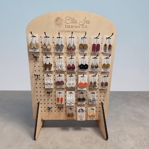 Könnte beinhalten: Ein hölzerner Ohrring-Präsentationsständer mit verschiedenen Ohrringpaaren. Der Ständer hat eine helle Holzoberfläche und trägt den Text "Ellie Joe Design Co." und "custombymonica.com". Die Ohrringe sind in verschiedenen Formen und Farben.