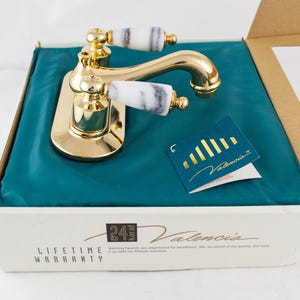 May include: A gold-colored faucet with white and gray marbled handles, presented in an open box with a teal lining. A small tag with gold text reads "Valencia". The box also displays the text "24 karat" and "Lifetime Warranty".