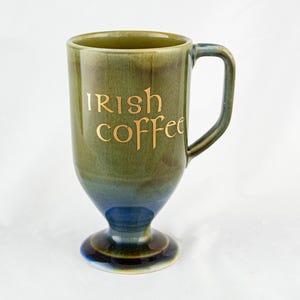 May include: A tall, green and blue Irish coffee mug with a handle. The words "Irish coffee" are written in gold lettering on the front of the mug. The mug has a pedestal base.