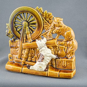 May include: A ceramic wall planter featuring a brown cat and a white dog figurine. The planter is shaped like a spinning wheel with a brown and green glaze. The planter has the text "Rus" on the bottom.