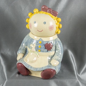 May include: A ceramic doll-shaped cookie jar with a lid. The jar features a light skin tone, yellow curly hair, and a pink bow. It wears a blue dress with a white apron and a heart-shaped patch. The jar is approximately 10 inches tall.