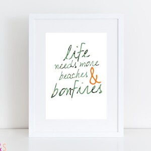 Op de afbeelding: Wit ingelijste kunstdruk met de woorden "life needs more beaches & bonfires" in groene en oranje letters. De woorden staan op een witte achtergrond. De print zit in een witte lijst.