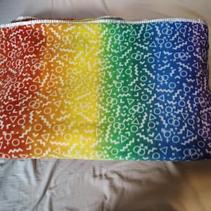 Könnte beinhalten: Eine gefaltete Decke mit einem Regenbogen-Farbverlauf, der von Rot nach Blau übergeht. Die Decke weist ein sich wiederholendes Muster aus weißen Geschlechtssymbolen, Dreiecken und Wellenlinien auf. Die Kanten sind mit einem silbernen Rand versehen.