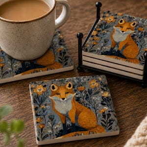 May include: A set of square ceramic coasters featuring a fox illustration surrounded by yellow flowers and blue foliage. The set includes a black metal holder, with one coaster placed under a speckled beige mug and another lying flat.