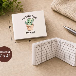 May include: A white pocket-sized notebook measuring 3 inches by 4 inches. The cover features a cartoon cactus in a pot with the text My Little Book Of Pricks. The interior pages contain a printed grid with columns labeled Date and Level.