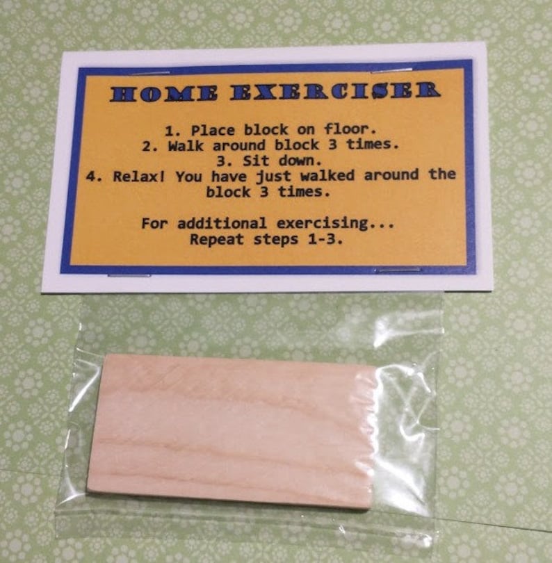 May include: A rectangular wooden block enclosed in a clear plastic bag, accompanied by a white card with a blue border and yellow center. The card features the text: HOME EXERCISER, 1. Place block on floor. 2. Walk around block 3 times. 3. Sit down.