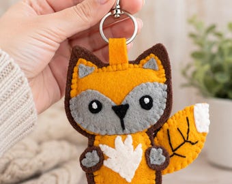 May include: A felt fox keychain featuring a bright orange body with dark brown edges, a gray face mask, black eyes, and a white chest patch. The fox has a bushy tail with a white tip and black stitched details, attached to a silver metal lobster clasp.