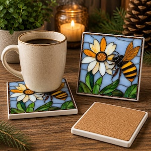 May include: A square ceramic coaster featuring a stained glass style design of a yellow and black bee on a white daisy with green leaves against a light blue background. The coaster has a cork backing and a white border.