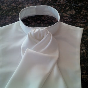 May include: A white satin fabric collar with a large, folded bow tie knot. The collar is designed to be worn with a formal shirt.
