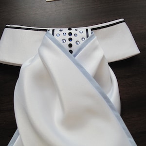 May include: White satin cravat with a black and light blue trim. The cravat has a cluster of rhinestones in the center.