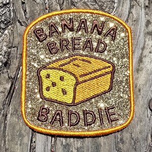 May include: A rectangular patch with a gold glitter background and a gold border. The patch features the words "BANANA BREAD BADDIE" in brown letters and an embroidered loaf of banana bread.