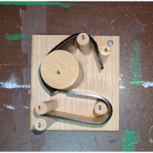 May include: A wooden model on a light-coloured wooden base. It features cylindrical wooden dowels, a large circular wooden piece, and a curved metal strip. The components are arranged to demonstrate a mechanical concept, possibly a pulley or similar device.