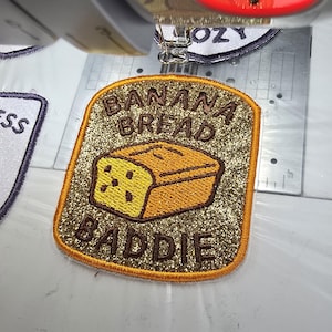 May include: A gold glitter patch featuring the text "BANANA BREAD BADDIE" above and below a cartoon banana bread loaf. The bread is yellow and brown, with a dark brown outline. The patch has a gold border.