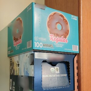 May include: A stack of three boxes of coffee pods. The top box is blue and features a donut graphic with the text "Regular Medium Roast Coffee". The middle box is blue and white with a mountain scene. The bottom box is blue and features the text "Organic and Fair Trade Certified".