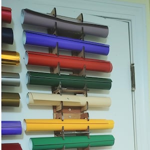 Pode incluir: Um suporte de madeira com 10 rolos de vinil em várias cores, incluindo roxo, vermelho, verde, amarelo e bege. O suporte está montado em uma porta branca.