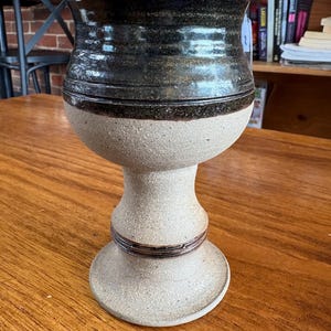 May include: A handcrafted ceramic chalice. It features a dark brown rim and a light tan body. The chalice has a pedestal base and a decorative band around the stem. The interior is dark brown. It sits on a wooden table.