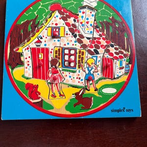 May include: A vintage wooden puzzle depicting a colourful house scene with two children, a squirrel, and a rabbit. The house has a red roof with polka dots, red doors, and a blue window. The puzzle is set against a blue background with a green and brown border. The text "simplex toys" is visible.