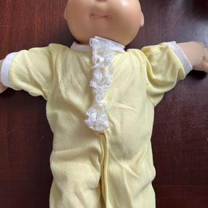 May include: A vintage Cabbage Patch Kid doll with a soft, vinyl head and a yellow onesie. The doll has blue eyes and a smiling expression. The onesie has white lace trim down the front and around the collar and cuffs.