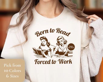 May include: A white t-shirt with brown text that reads "Born to Read Forced to Work". The text is surrounded by a vintage-style illustration of two women reading and working.