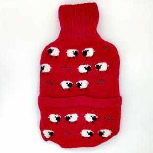 May include: A red knitted hot water bottle cover featuring a pattern of white sheep with black faces and legs. The cover has a turtleneck-style opening and a folded bottom edge. Small green accents are scattered throughout the design.