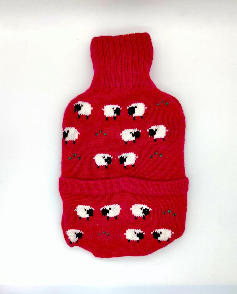 May include: A red knitted hot water bottle cover featuring a pattern of white sheep with black faces and legs. The cover has a turtleneck-style opening and a folded bottom edge. Small green accents are scattered throughout the design.