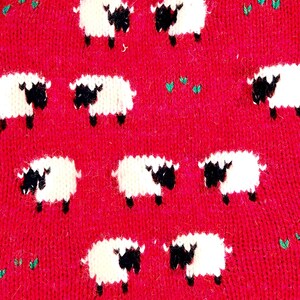 May include: Close-up of a red textile with a repeating pattern of white sheep with black faces and legs. Small green leaf-like shapes are scattered throughout the design. The sheep are evenly spaced across the fabric.
