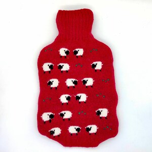May include: A red knitted hot water bottle cover featuring a pattern of white sheep with black faces and legs. The cover has a ribbed neck and is designed to fit a standard hot water bottle. Green accents are scattered throughout the design.
