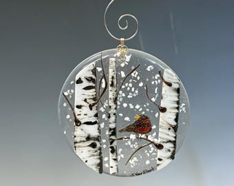 May include: A round glass ornament featuring a winter scene with birch trees and a red bird. The trees have black and white bark, and the background is a snowy gray. A silver hook is attached for hanging.