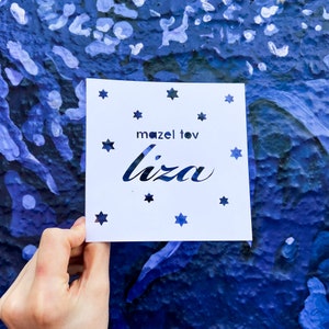 May include: A white card with the text "mazel tov Liza" in black cursive lettering. The card is decorated with small, cut-out stars.