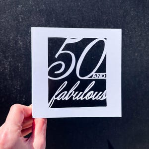 Puede incluir: Tarjeta blanca con fondo negro y el texto "50 and fabulous" en letra cursiva blanca.