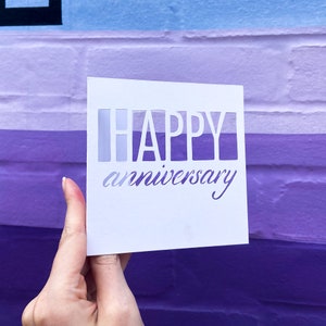 Puede incluir: Una tarjeta blanca con las palabras "HAPPY anniversary" escritas en letras blancas sobre un fondo de pared de ladrillo morado y azul.