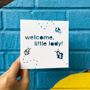 Puede incluir: Una tarjeta cuadrada blanca con las palabras "welcome, little lady!" recortadas en color turquesa. La tarjeta presenta diseños de mariquitas turquesas y líneas punteadas. La tarjeta se sostiene frente a una pared de ladrillos azules.