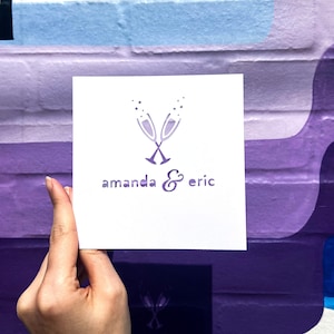 Puede incluir: Una tarjeta cuadrada blanca con un diseño púrpura de dos copas de champán y los nombres "amanda & eric" en una fuente estilizada. La tarjeta se sostiene contra una colorida pared pintada de púrpura y azul.