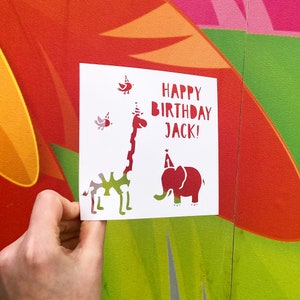 Puede incluir: Una tarjeta blanca con una jirafa roja, un elefante y pájaros. La tarjeta dice "¡Feliz cumpleaños Jack!"
