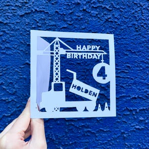 Puede incluir: Tarjeta de papel recortada en blanco con un diseño de grúa de construcción. La tarjeta dice "Feliz cumpleaños" y "Holden" con el número "4" en un círculo.