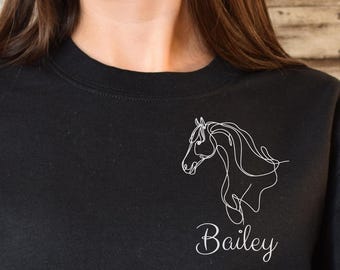 May include: Black sweatshirt featuring a white line drawing of a horse's head and the name "Bailey" in cursive. The sweatshirt is a crew neck style and appears to be made of a soft material. The person is wearing blue jeans.