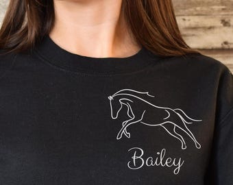 May include: Black sweatshirt featuring a white outline of a horse in motion, with the name "Bailey" in cursive script below. The long-sleeved top is made of a soft material and is suitable for casual wear.