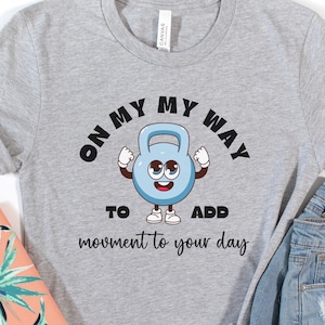 Puede incluir: Una camiseta gris con el texto "ON MY WAY TO ADD movement to your day" y un personaje de dibujos animados de pesa rusa. La imagen incluye pantalones cortos de mezclilla y un clutch con estampado coral y turquesa.
