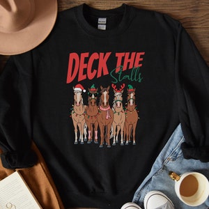 May include: Black sweatshirt with a Christmas-themed design featuring four horses wearing Santa hats and elf hats. The text "Deck the Stalls" is printed in red on the sweatshirt.