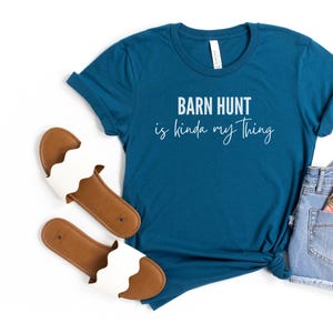 May include: Teal blue t-shirt with the text "Barn Hunt is kinda my thing" in white script font. The shirt is paired with white sandals and light blue denim shorts.